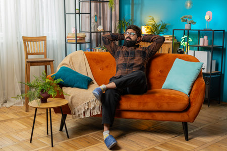 Bearded man in living room relaxing on sofa closed his eyes. Happy positive guy taking a break resting, puts hands behind head sitting at home couch. Stress free peaceful dayの写真素材
