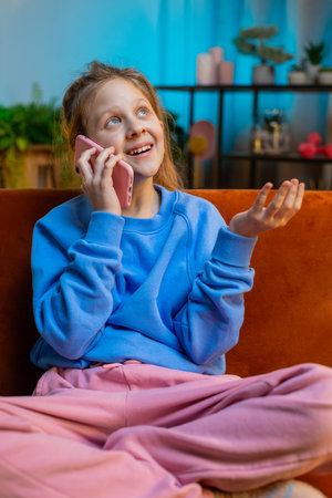 Phone call, good news, gossip. Happy surprised preteen child girl in pleasant conversation on smartphone, enjoying talking with friend. Female kid at home night living room apartment on sofa. Verticalの写真素材