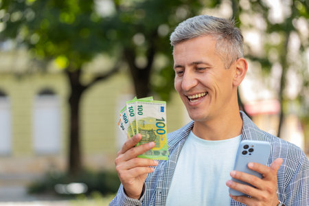 Rich man counting holding money euro cash, using smartphone calculator app, plans to order gifts and food delivery online, booking hotel room. Gray-haired guy tourist in city street, outdoors.の写真素材