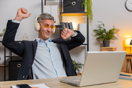 Happy relaxed young businessman working on laptop at office wearing headphones and sunglasses listening favorite energetic disco music, dancing. Freelancer man relaxing, taking a break.の写真素材