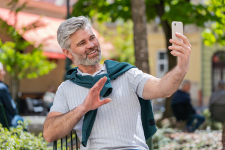 Bearded mature man blogger using smartphone, communicating video call online with subscribers,...