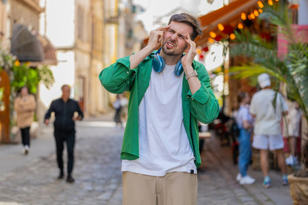 Displeased exhausted Caucasian man rubbing temples to cure headache problem suffering from tension migraine stress. Young guy tourist grimacing, pain high blood pressure standing on city urban street.の写真素材