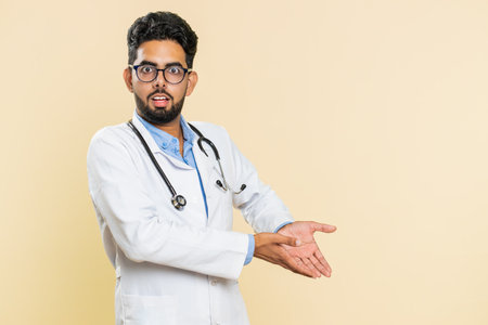 Indian young doctor cardiologist man raising hands asking what why reason of failure, demonstrating disbelief irritation by troubles. Isolated on beige backgroundの写真素材