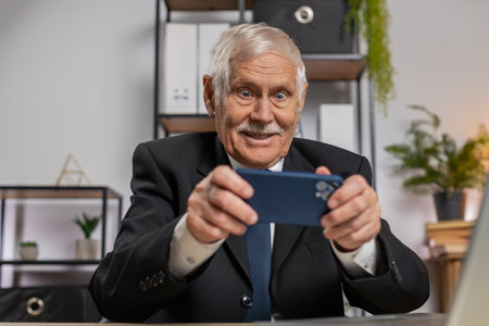 Caucasian senior businessman playing on smartphone exciting mobile application celebrate winning racing, shooter game at office desk workplace. Professional freelancer man enjoy good work of new app.の写真素材