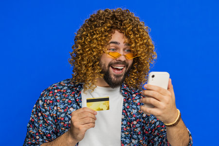 Happy excited man using credit card and smartphone while transferring spending money, purchases online shopping, ordering food delivery. Stylish guy isolated on blue background indoorsの写真素材
