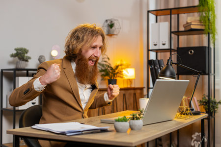 Caucasian bearded businessman working on laptop shocked by sudden victory, game winning goal achievement, good career news. Excited happy joyful guy freelancer clenching fists at home office.の写真素材