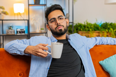 Handsome tired smiling man drinking cup of warm coffee or herbal tea in the morning. Guy enjoying refreshment comfortable relaxing alone sitting in home apartment room on orange sofa.の写真素材