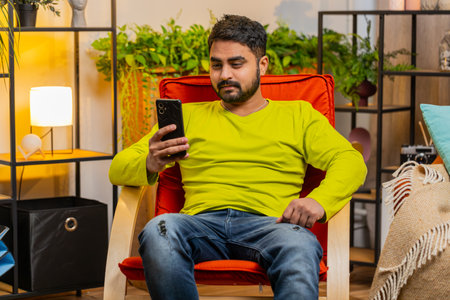 Man sits on chair uses mobile phone smiles at home room apartment. Young guy texting share messages...