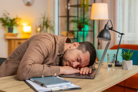 Bored sleepy young businessman working on laptop, sleeping on netbook at home office desk in living...