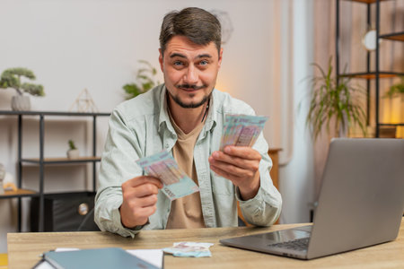 Planning budget. Rich happy man counting money Ukrainian hryvnias UAH cash using laptop calculate...