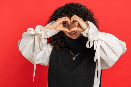 Woman in love. Smiling attractive Caucasian young woman with curly hair makes heart gesture demonstrates love sign expresses good positive feelings and sympathy. Girl isolated on red backgroundの写真素材