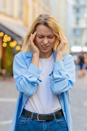 Displeased exhausted Caucasian woman rubbing temples to cure headache problem suffering from tension migraine stress. Young lady tourist grimacing pain high blood pressure stands on city urban streetの写真素材