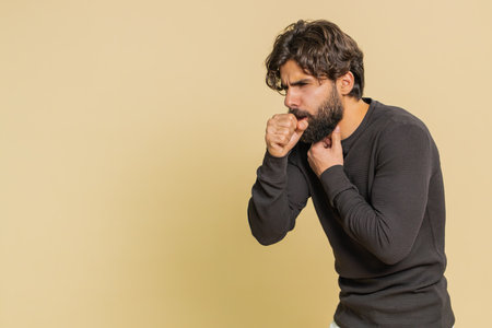 Young man coughing covering mouth with hand feeling sick, allergy or viral infection disease symptoms, infection, pain, virus. Guy isolated on beige studio backgroundの写真素材