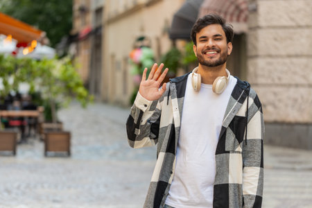 Indian young man smiling friendly at camera, waving hands gesturing hi, greeting or goodbye, welcoming with hospitable expression outdoors. Arabian guy tourist in urban city street. Lifestyles.の写真素材