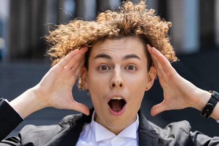 Caucasian businessman looking at camera shocked by sudden victory, game winning, lottery goal achievement, good news. Professional manager man with curly brown hair outside office.の写真素材