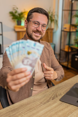 Young businessman freelancer holding fan of cash money Ukrainian hryvnias UAH banknotes, pointing and showing thumbs up, celebrate, success career, online income, wealth in living room at home office.の写真素材