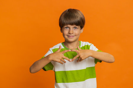 I love you. Smiling adorable young preteen child boy kid makes heart gesture demonstrates love sign expresses good positive feelings and sympathy. Happy little children isolated on orange backgroundの写真素材