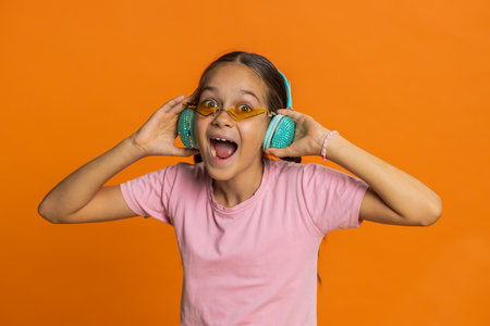 Happy smiling preteen child girl in headphones listening music dancing disco fooling around having fun expressive gesticulating hands relaxing on party making funny moves. Kid on orange backgroundの写真素材