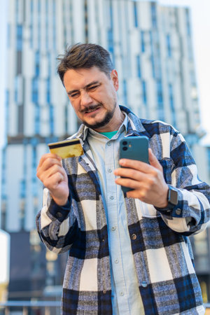 Frustrated businessman discovered fraud while checking credit card on mobile phone standing on downtown city street. Sad man tourist became a victim of deception, money had been stolen from account.の写真素材