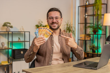 Young businessman freelancer holding fan of cash money euro banknotes, pointing and showing thumbs up, celebrate, success career, online income, wealth in living room at home office desk apartment.の写真素材