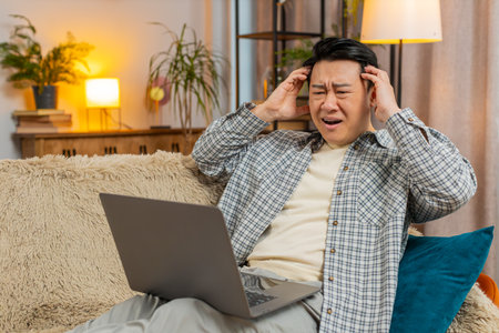 Upset displeased man with brown hair using laptop sitting on sofa in living room at home apartment....