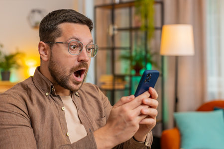 Caucasian young businessman using smartphone shocked by sudden victory game winning, lottery goal achievement, good news. Professional manager freelancer guy sitting in home office.の写真素材