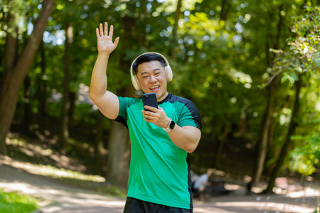 Athletic fitness adult guy sport runner man walking, wearing headphones listening music from smartphone and smiling. Healthy sportsman workout cardio outdoors in morning park on summer day.の写真素材