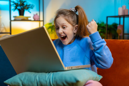 Caucasian teenager girl child kid use laptop computer, receive good news message, shocked by sudden victory, celebrate game play win, lottery jackpot, purchases online shopping at evening home roomの写真素材