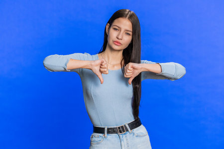 Dislike. Upset unhappy young woman showing thumbs down sign gesture, expressing discontent, disapproval, dissatisfied, dislike feedback. Pretty girl. Indoors isolated on blue studio wall backgroundの写真素材