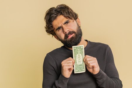 Poor young man with insufficient amount of money, holding one dollar banknote. Financial crisis. Bankruptcy. Poverty and destitution. Adult guy standing on beige background.の写真素材