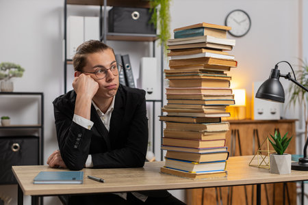 Tired exhausted stressed young businessman looking at large pile of books and looking at volume of...