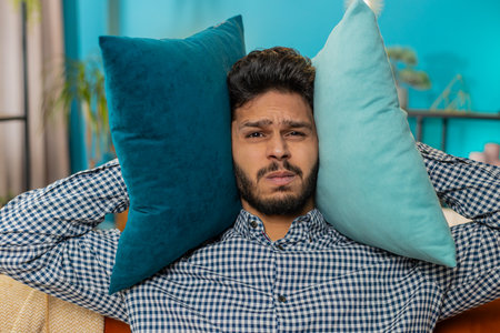 Repair work at neighbors. Irritated young man cover ears with pillows annoyed by noisy loud music...