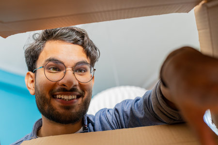 View from inside the box. Happy man unpacking delivery parcel at home. Smiling satisfied guy shopper online shop customer opens cardboard box receive purchase gift by fast postal shippingの写真素材