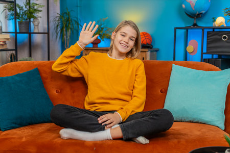 Caucasian teenager boy waves with a smile while greeting happily at home. Sits on the sofa, looking...