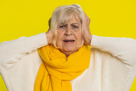 Frustrated annoyed irritated senior old woman covering ears gesturing no avoiding advice ignoring unpleasant noise loud voices. Mature grandmother on yellow backgroundの写真素材