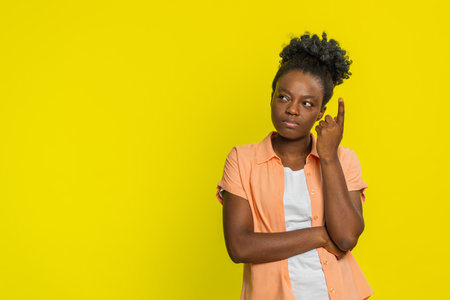 African American woman touches temple with finger, showing deep thought and focused expression. Black girl isolated on yellow background, reflecting concentration and search for creative solutionsの写真素材
