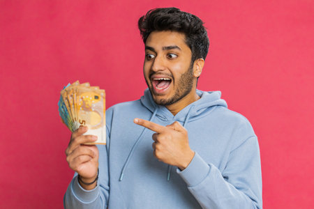 Happy man holding euro cash, counting money with satisfaction, showing profit, salary, or bank loan success. Excited guy displaying wealth and motivation, isolated on red backgroundの写真素材