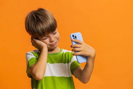 Upset sad preteen child boy kid use smartphone typing browsing, play game, loses, surprised by sudden results. Sad children on orange background indoorsの写真素材