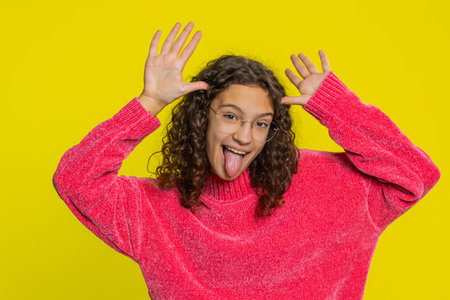 Caucasian teenager girl in blouse showing tongue making faces at camera, fooling around, joking, aping with silly face. Young teen child isolated on yellow background indoorsの写真素材