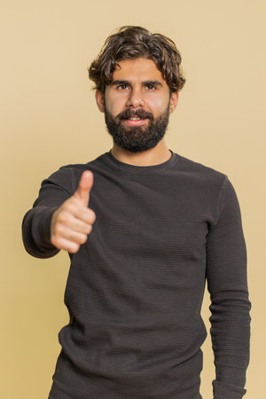 Happy man in long sleeve raises thumbs up agrees with something, gives positive reply recommends advertisement likes good. Bearded guy isolated alone on beige studio background.の写真素材