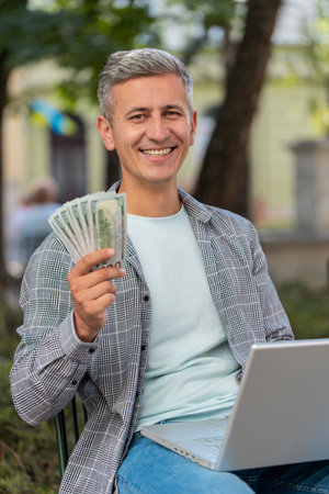 Happy rich man using laptop winner victory counting holding fan of cash money in dollar banknotes sitting on chair city street park. Young male guy tourist planning budget online shopping. Verticalの写真素材