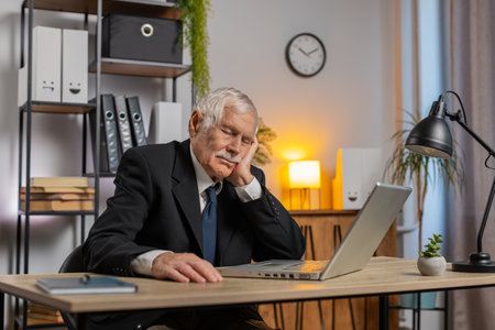 Bored sleepy elderly businessman working on laptop computer, leaning on hand at home office desk. Exhausted tired grandfather freelancer workaholic guy falling asleep. Employment, occupation, worklessの写真素材