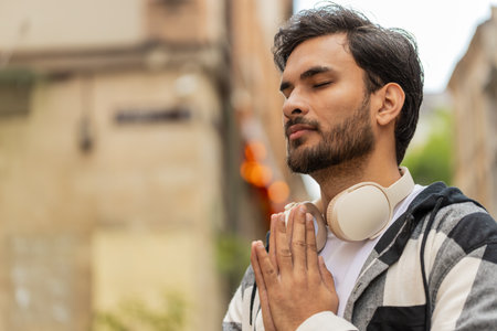 Man praying with closed eyes outdoors. Young guy in casuals clasping hands in urban city street. Town lifestyleの写真素材