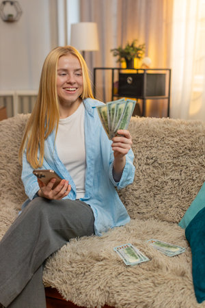 Young woman relaxes at home, counting banknotes while entering sums on smartphone to find extra savings. Caucasian girl on sofa celebrates with triumph pose, feeling proud of unexpected financial gainの写真素材