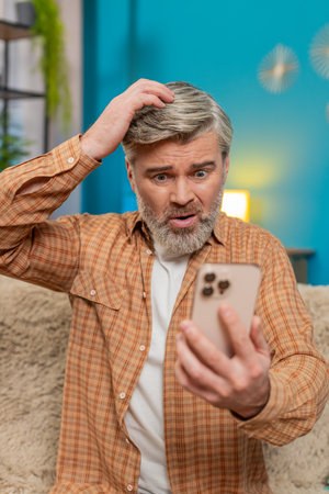 Mature man sitting on sofa at home, holding smartphone and showing signs of stress. Middle-aged guy...