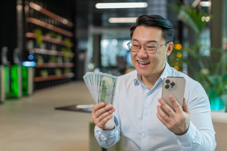 Successful happy rich mature businessman in shirt holding smartphone and counting dollar cash bill money fan sitting in office lobby. Happy Chinese guy winning online lottery game. Calculating budget.の写真素材