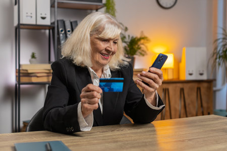 Happy Caucasian senior woman using bank credit card and smartphone for online shopping payments at...