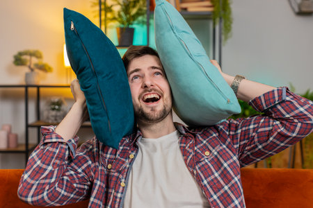 Irritated Caucasian tired man annoyed by noisy neighbors suffers from headache on couch. Thin walls at home, flat without sound insulation, repair work concept. Guy covers ear with cushion pillows.の写真素材