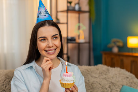 Young woman at home joyfully celebrating birthday holding cake with lit candles smiling happily....