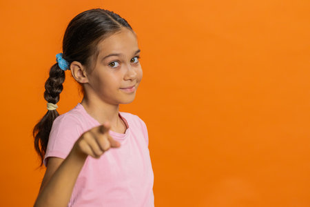 Happy young preteen child girl kid pointing finger forward to camera, choosing lucky game winner, indicating to awesome you, promotion. Smiling teenager children isolated on orange backgroundの写真素材